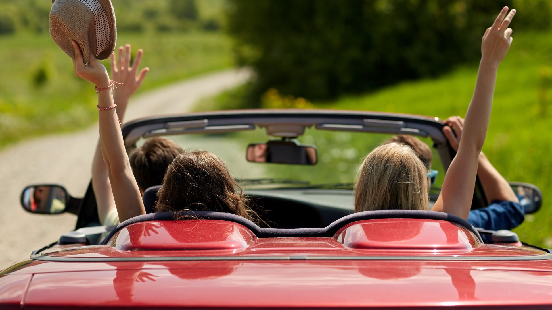 Group of friends driving in a red convertible car and holding their hands up in the air.